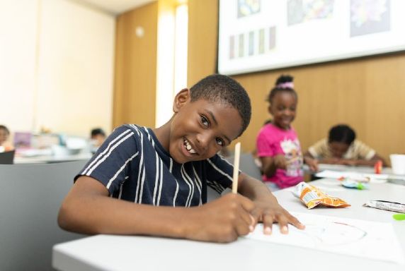 A young boy smiling at the camera while sitting and drawing at a table. Other children are at the table in the background.