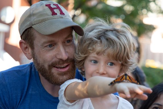 A father and child gazing in wonder at a monarch butterfly resting on the child's outstretched right hand.