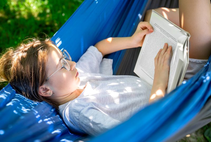 A girl reclining outdoors in a blue hammock, reading a book.