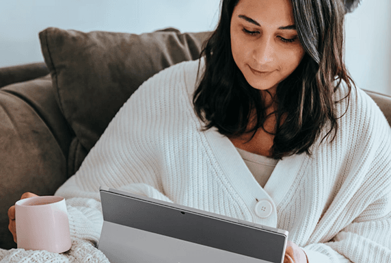 A person sits on a couch while reading on a tablet. They are holding a mug in one hand and the tablet in the other.